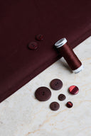 Tissu coton déperlant bordeaux avec un fuseau de fil assorti et des boutons ton sur ton, dont un bouton rouge nacré, posés dessus.