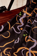 Tissu viscose Misty noir à motif graphique de volutes lilas et moutarde, drapé sur l'accoudoir en bois d'une chaise vintage.