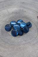 Boutons ronds bombés à queue, sans trous apparents, finition nacrée bleu ardoise, présentés en tas sur une pierre grise, avec un brin d'herbe séchée en arrière-plan.
