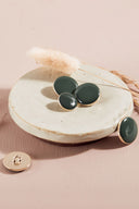 Boutons ronds et ovales bombés, finition brillante, coloris vert cèdre grisé, dont un présenté de dos laissant voir l'attache à queue métallique, sur une coupelle en céramique blanche, fond rose avec un épi de lagurus séché.