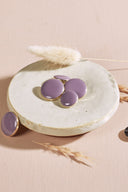 Quatre boutons ronds et ovales bombés, finition brillante, coloris parme mauve pâle, cerclés d'un fin liseré doré, présentés sur une petite coupelle en céramique blanche, sur fond rose avec un épi de lagurus séché.
