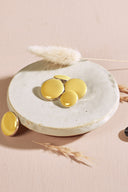 Quatre boutons ronds et ovales bombés, finition brillante, coloris jaune citron, cerclés d'un fin liseré doré, présentés sur une petite coupelle en céramique blanche, sur fond rose avec un épi de lagurus séché.