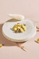 Boutons ronds bombés jaune citron cerclés d'un fin liseré doré, à queue métallique (shank), présentés en tas dans une petite coupelle en céramique blanche sur fond rose, avec un brin d'herbe séchée (lagurus) en arrière-plan.