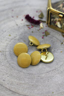 Cinq boutons ronds facettés moutarde cerclés d'un liseré doré, à queue métallique (shank), posés sur une pierre grise avec des fleurs séchées et un coffret doré flou en arrière-plan.