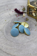 Cinq boutons ronds facettés bleu gris clair (storm) cerclés d'un liseré doré, avec un anneau de couture doré visible, sur pierre grise avec fleurs séchées et coffret doré flou.