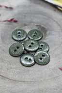 Boutons ronds à deux trous entièrement pailletés vert kaki, présentés en tas sur une pierre grise, avec un fil de raphia bordeaux en arrière-plan.