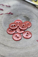 Boutons ronds à deux trous entièrement pailletés rouge terracotta, présentés en tas sur une pierre grise, avec un fil de raphia bordeaux en arrière-plan.