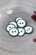 Boutons ronds à deux trous en nacre blanche, cerclés d'un liseré pailleté vert cactus, présentés en tas dans une coupelle en pierre grise, avec des pétales de fleurs blanches et bordeaux en arrière-plan.