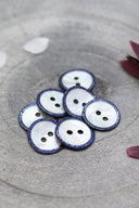 Boutons ronds à deux trous en nacre blanche, cerclés d'un liseré pailleté bleu cobalt, présentés en tas dans une coupelle en pierre grise, avec des pétales de fleurs blanches et bordeaux en arrière-plan.