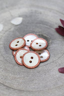 Boutons ronds à deux trous en nacre blanche, cerclés d'un liseré pailleté rose cuivré, présentés en tas dans une coupelle en pierre grise, avec des pétales de fleurs blanches et bordeaux en arrière-plan.