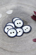 Boutons ronds à deux trous en nacre blanche, cerclés d'un liseré pailleté bleu marine, présentés en tas dans une coupelle en pierre grise, avec des pétales de fleurs blanches et bordeaux en arrière-plan.