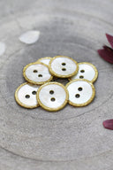 Boutons ronds à deux trous en nacre blanche, cerclés d'un liseré pailleté or moutarde, présentés en tas dans une coupelle en pierre grise, avec des pétales de fleurs blanches et bordeaux en arrière-plan.