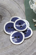 Boutons ronds à deux trous, finition émaillée bleu cobalt cerclée d'un liseré blanc nacré, présentés en tas sur une pierre grise, avec une feuille verte et une petite coupelle en verre facetté en arrière-plan.
