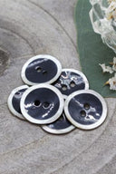 Boutons ronds à deux trous, finition émaillée gris anthracite cerclée d'un liseré blanc nacré, présentés en tas sur une pierre grise, avec une feuille verte et une petite coupelle en verre facetté en arrière-plan.