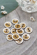Boutons ronds à deux trous, finition émaillée ocre cerclée d'un liseré blanc nacré, présentés en tas sur une feuille verte, avec une petite coupelle en verre facetté et des pétales de fleurs blanches en arrière-plan.