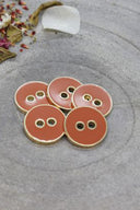 Cinq boutons ronds à deux trous, finition émaillée brillante orange terracotta chaud, cerclés d'un liseré métallique doré, posés sur une pierre grise avec des pétales de fleurs séchées éparpillés en arrière-plan.