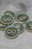 Sept petits boutons ronds à deux trous, finition pailletée dorée sur fond vert cèdre foncé, cerclés d'un liseré métallique doré, photographiés en gros plan sur une pierre grise.
