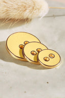 Trois boutons ronds à deux trous de tailles dégradées, finition émaillée brillante jaune citron doré, cerclés d'un liseré métallique doré, posés sur une pierre claire avec un brin d'herbe séchée (lagurus) en arrière-plan.