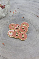 Sept boutons ronds à deux trous, finition émaillée brillante rose pêche melba, cerclés d'un liseré métallique doré, posés sur une pierre grise, avec un petit vase transparent et des pétales séchés en arrière-plan.