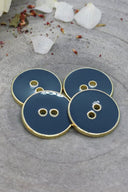 Cinq boutons ronds à deux trous, finition émaillée brillante bleu ardoise gris, cerclés d'un liseré métallique doré, posés sur une pierre grise avec des pétales de fleurs séchées éparpillés en arrière-plan.