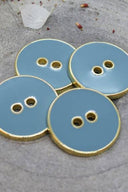 Quatre boutons ronds à deux trous, finition émaillée brillante bleu-gris clair orageux, cerclés d'un liseré métallique doré, posés sur une pierre grise avec des pétales de fleurs séchées éparpillés en arrière-plan.