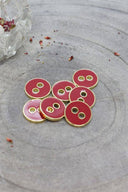 Cinq boutons ronds à deux trous, finition émaillée brillante rouge terracotta, cerclés d'un liseré métallique doré, posés sur une pierre grise avec des pétales de fleurs séchées éparpillés en arrière-plan.