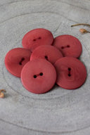Sept boutons ronds classiques à deux trous, effet bois veiné rouge terracotta, présentés en tas sur une pierre grise, avec une feuille séchée en arrière-plan.