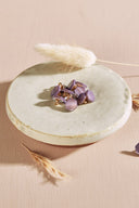 Boutons de forme irrégulière façon pierre facettée, coloris mauve parme, avec fixation à queue métallique dorée, présentés en tas dans une petite coupelle en céramique blanche sur fond rose, avec un brin d'herbe séchée (lagurus) en arrière-plan.