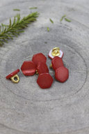 Boutons de forme irrégulière façon pierre facettée, coloris terracotta rouge brique, avec fixation à queue métallique dorée, posés sur une pierre grise avec un brin de romarin en arrière-plan.