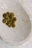 Une douzaine de petits boutons ronds brillants à deux trous, coloris vert olive kaki (dreamy twilight), présentés en tas dans une coupelle en pierre beige marbrée.