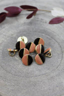 Two-tone round buttons with a gold metal shank, half black and half peachy melba pink, separated by a thin gold trim, piled on grey stone with a sprig of burgundy foliage in the background.