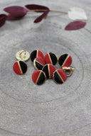 Boutons ronds bicolores à queue métallique dorée, moitié noire et moitié rouge terracotta, séparés par un fin liseré doré, présentés en tas sur une pierre grise avec un rameau de feuillage bordeaux en arrière-plan.