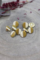 Boutons ronds bicolores à queue métallique dorée, moitié blanc cassé et moitié jaune moutarde, séparés par un fin liseré doré, présentés en tas sur une pierre grise avec un rameau de fleurs séchées bordeaux en arrière-plan.