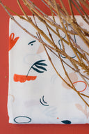 Tissu Sandstorm écru à motif de formes abstraites orange et marine, plié devant un fond terracotta avec des brindilles séchées.