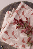 Tissu Windy rose poudré à motif graphique de feuilles courbes terracotta et écru, plié sur un socle en bois avec une branche de fleurs séchées bordeaux.