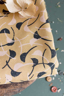 Tissu Windy moutarde à motif graphique de feuilles courbes marine et écru, plié sur un tabouret en bois avec une fleur séchée et des boutons gris, caramel et rose dispersés.