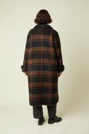 Patron Le Manteau – Femme de dos, manteau long tartan brun/noir, pantalon gris, derbies noires – Atelier Brunette