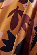 Tissu Seraphine moutarde à motif de feuilles graphiques anthracite, bordeaux et rose, drapé sur un tabouret en bois rond, avec des branches d'eucalyptus séchées.