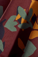 Tissu viscose Seraphine bordeaux à imprimé floral graphique moutarde et sauge, drapé sur un tabouret en bois rond avec des feuilles d'eucalyptus.