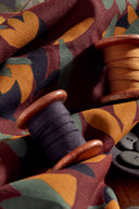 Tissu Seraphine bordeaux plié avec une bobine de fil moutarde en bois et des boutons gris-vert.